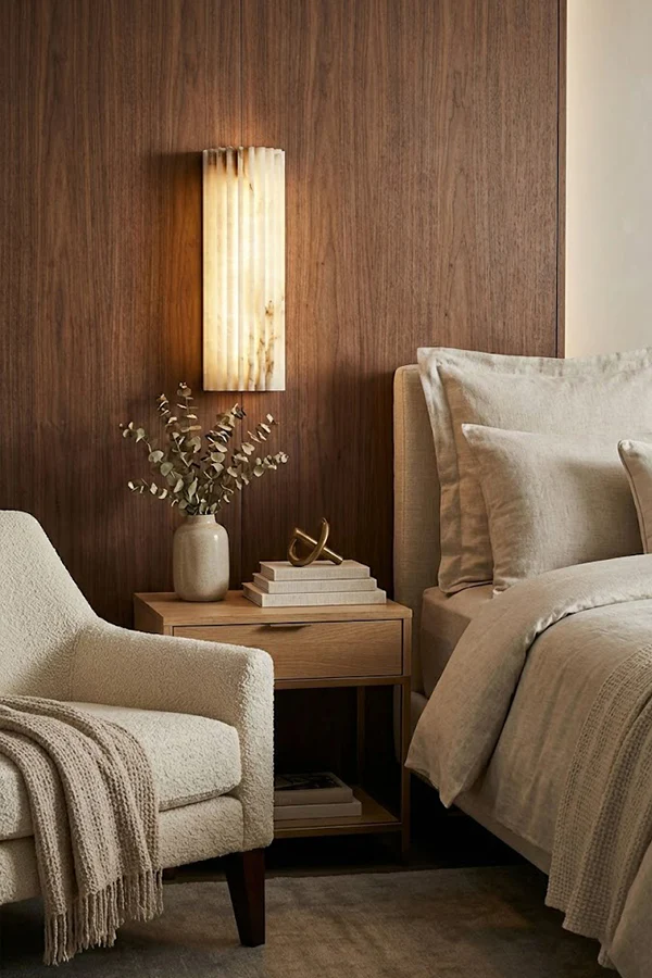 Aurelius Fluted Alabaster Sconce lit on dark wood panel wall, neutral cozy bedroom with bedside table and cream armchair