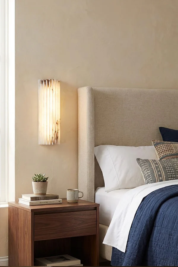 Aurelius Fluted Alabaster Sconce beside upholstered bed headboard, warm neutral bedroom with wooden nightstand and blue bedding