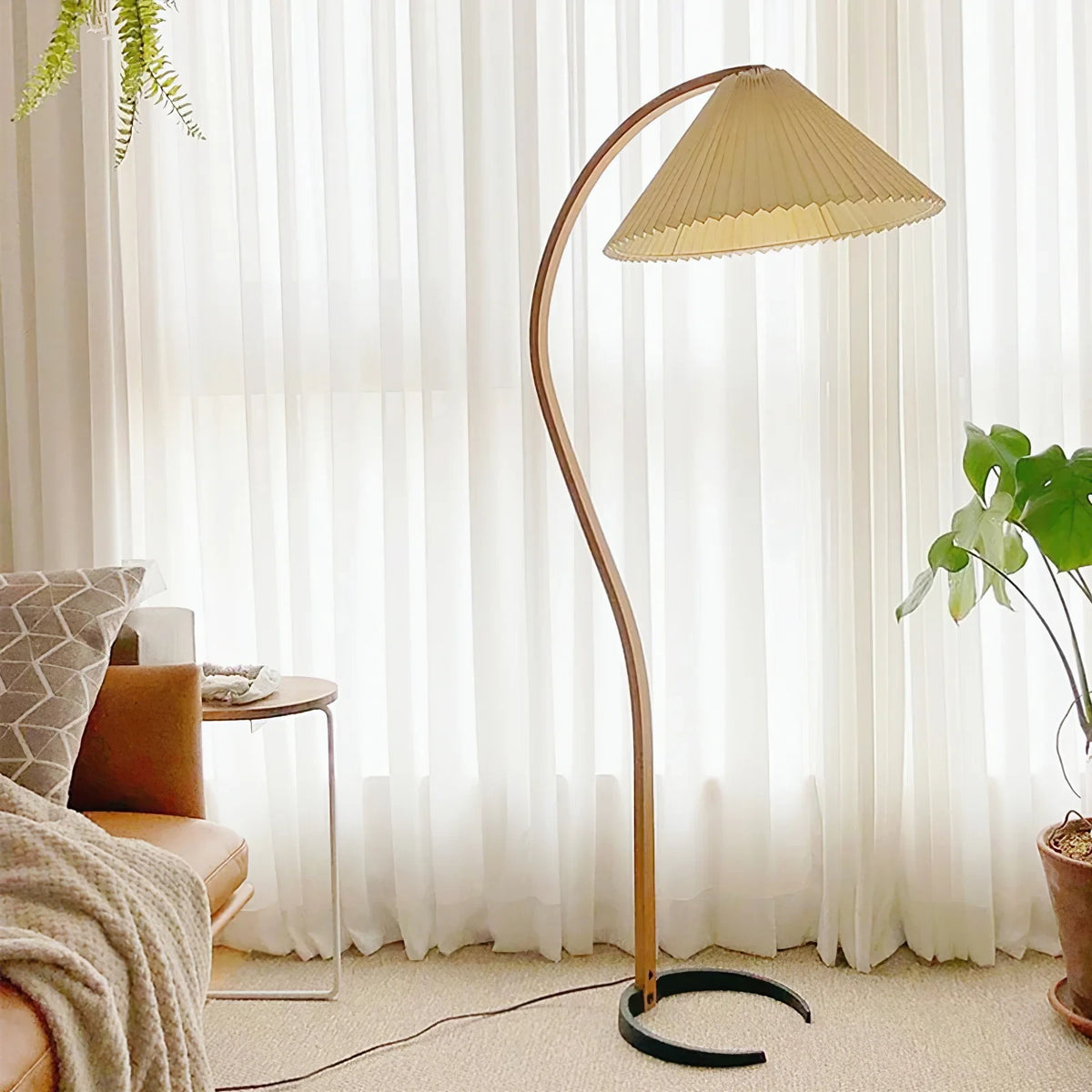 Caprani Floor Lamp with curved wooden frame and beige pleated shade, placed beside sofa in cozy living room with sheer curtains, interior lifestyle photo