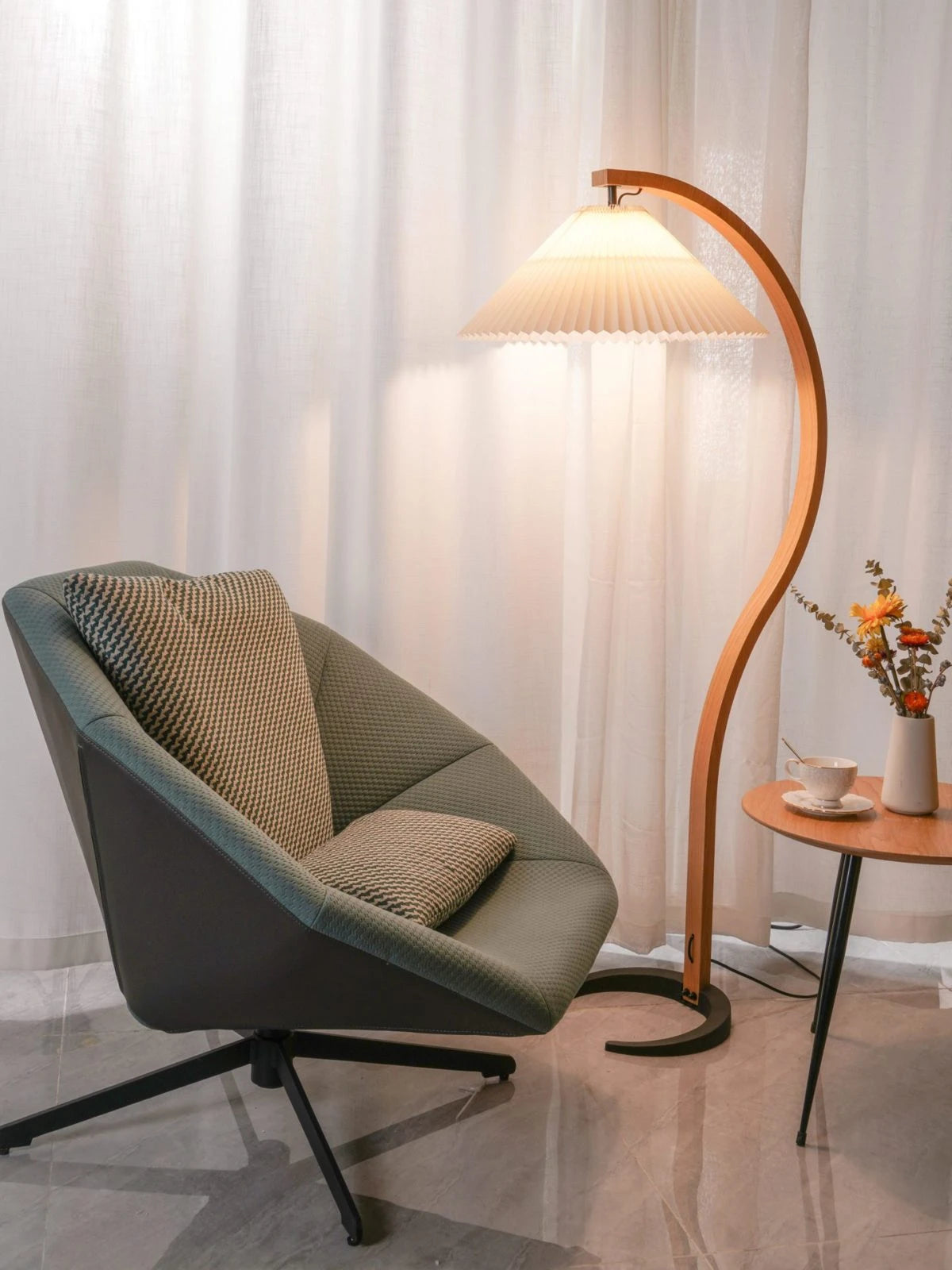Lit Caprani Floor Lamp curved wooden frame with illuminated cream pleated shade, arranged next to lounge chair in bright modern living room