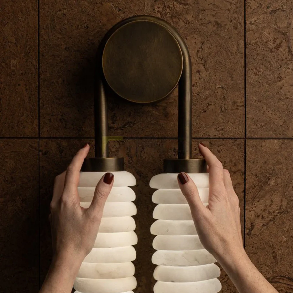 Hands adjusting twin alabaster sconce fixture on dark tiled bathroom wall